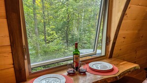 A dining table with plates, glasses, and a bottle of wine, set against a window overlooking a forest.