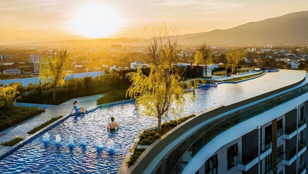 Ein Schwimmbecken mit Wasserspielen, umgeben von Bäumen und Gebäuden, bei Sonnenuntergang.