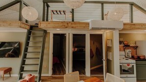 Un vaste espace de vie de style loft, doté d’un haut plafond, de poutres apparentes, d’un escalier, d’un coin repas avec des meubles en osier et d’une kitchenette.