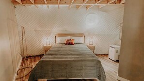 Une chambre compacte, composée d’une seule pièce, avec un plafond en bois, un lit recouvert d’une couverture verte, deux lampes de chevet et un petit radiateur.