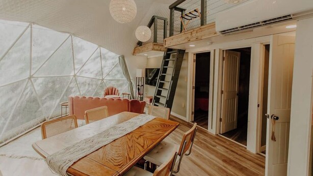 Une pièce spacieuse avec une grande table à manger en bois, un coin salon confortable et une vue dégagée grâce à une structure de fenêtre aux formes géométriques.