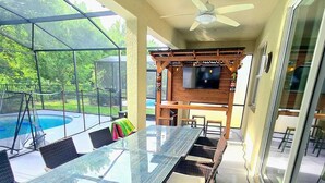 A covered outdoor dining area with a glass table, wicker chairs, a ceiling fan, and a TV screen.