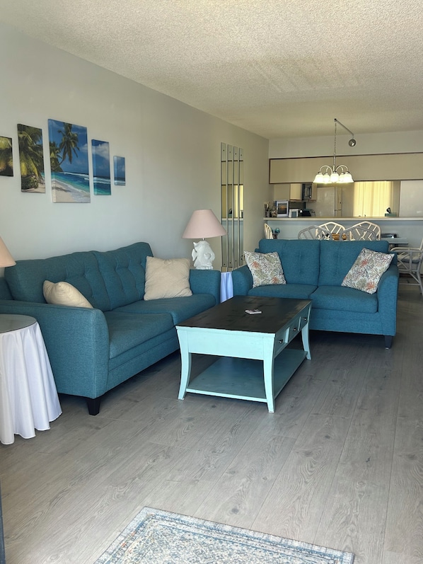 A living room with two blue sofas, a coffee table, and a small side table. There are wall-mounted pictures, a lamp, and a kitchen area in the background.