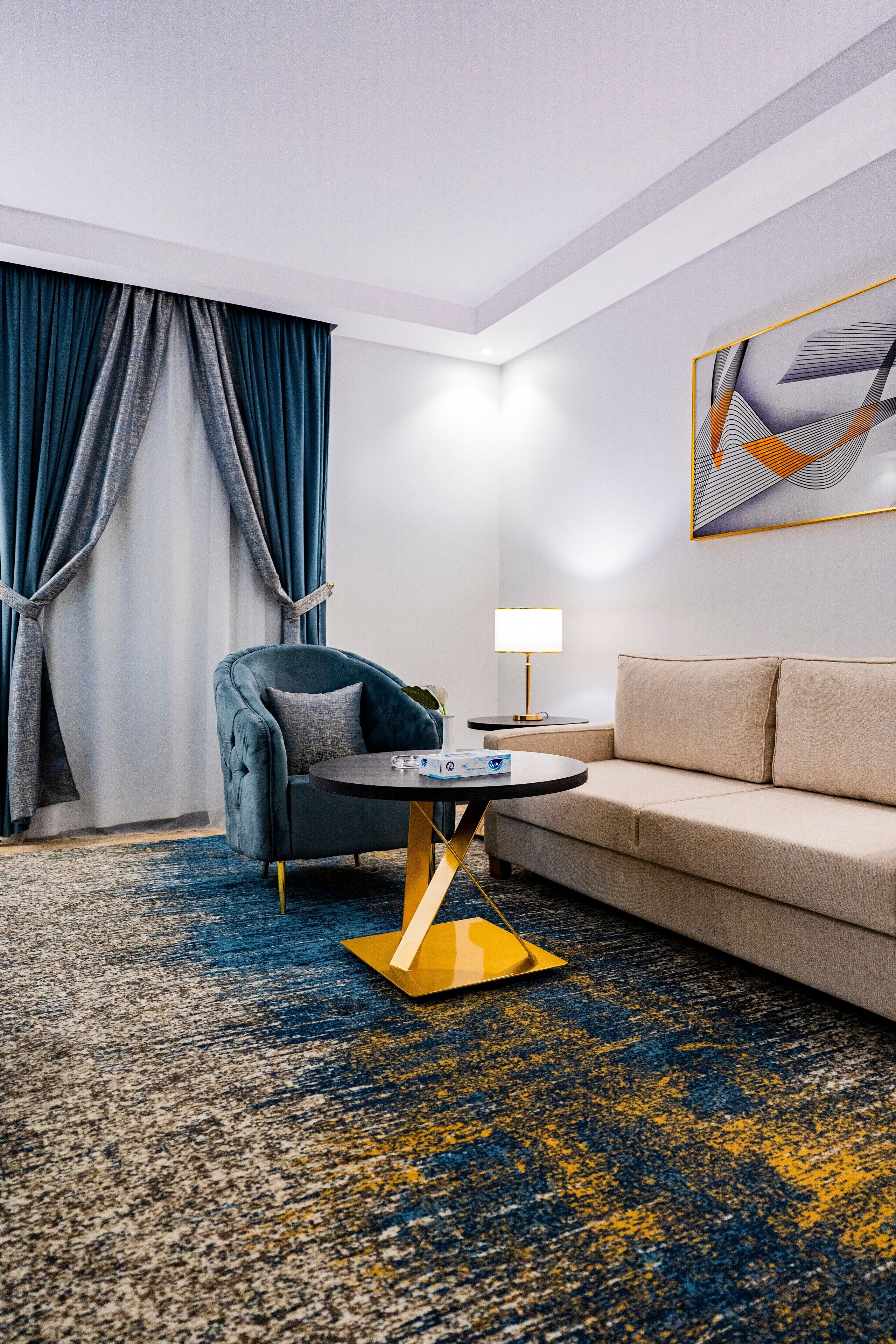 A modern hotel room with a blue armchair, a beige sofa, a round coffee table, and a patterned carpet.