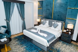 A modern hotel room with a large bed, two bedside tables, a nightstand, a blue and white patterned rug, and a teal armchair. - ODST Jed (Jeddah)