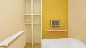 A small, narrow room with a bed, a white shelf, and a television mounted on the wall. - Backpacker Homestay Cibubur by PEKOTEL (Bekasi)