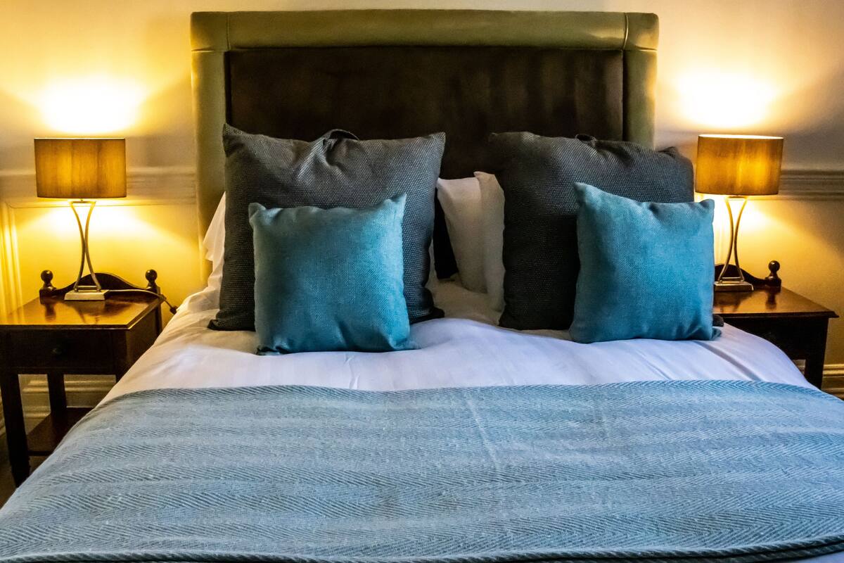 Een net opgemaakt bed met een blauwe deken en meerdere kussens, aan weerszijden geflankeerd door nachtkastjes met lampen.