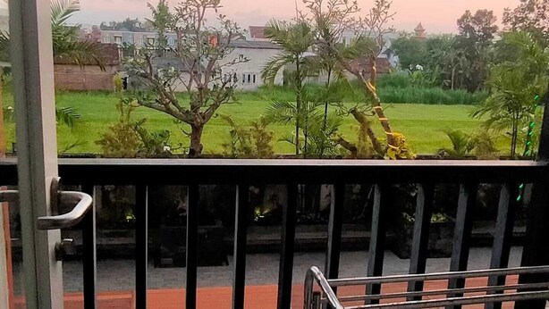 A view from a balcony with a swimming pool, a grassy area with trees, and a building in the background. - Student Castel By PEKOTEL (Depok)