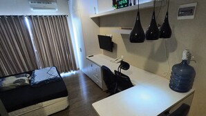 A compact hotel room with a bed, a desk, a chair, a water dispenser, and a clock on the wall. - Student Castel By PEKOTEL (Depok)