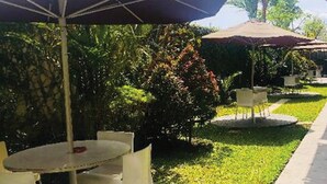 An outdoor seating area with tables and chairs under umbrellas, surrounded by greenery and a pathway. - Student Castel By PEKOTEL (Depok)