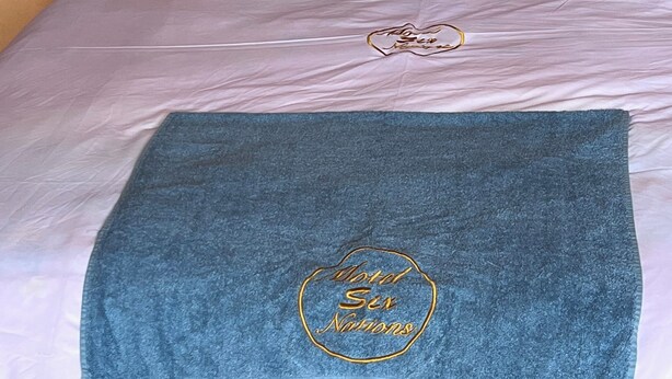 Cama com lençóis brancos e toalha azul com a inscrição 'Motel Six Nations'.