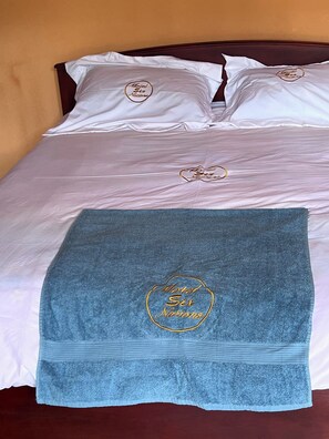 A bed with white linens and a blue towel with 'Motel Six Nations' embroidered on it. - Motel Six Nations (Boumnyébel)