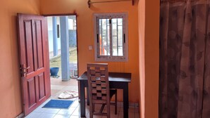 Quarto com porta de madeira, janela com cortina estampada com padrão de folha, uma pequena mesa e uma cadeira.