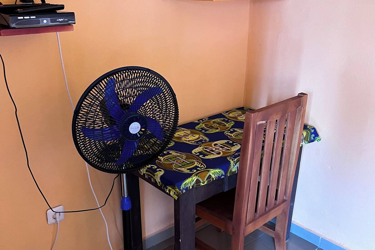 Une pièce d’angle avec une chaise en bois, un ventilateur sur pied, une étagère et un téléviseur fixé au mur.