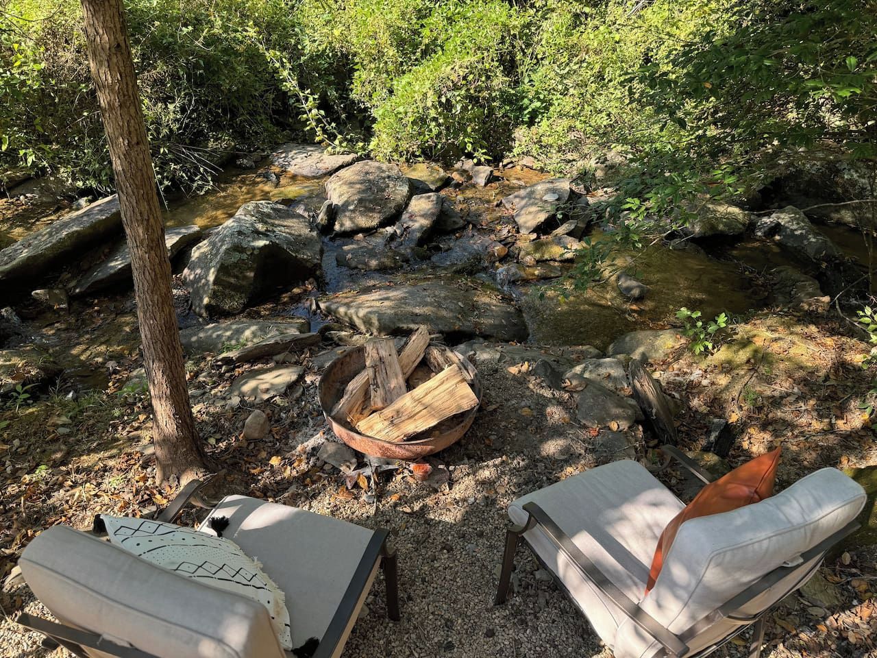A campsite with a fire pit surrounded by rocks, two camping chairs, and a small stream.