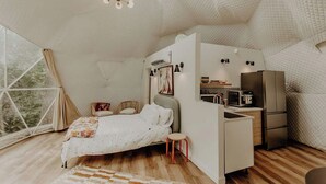 Ein kleines Schlafzimmer mit Bett, einem Hocker, einer Küchenzeile und einem Fenster mit Vorhängen.