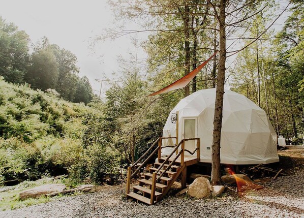 A geodesic dome tent with a wooden staircase leading up to a door, surrounded by trees and vegetation. - Whispering Waters: Dome Getaway (Cosby)