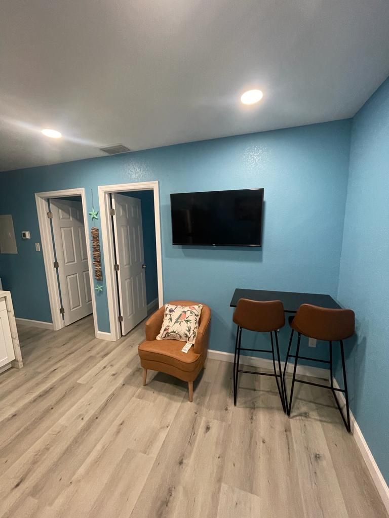 A room with a blue wall, a flat-screen TV mounted above a small table with two chairs, a brown armchair with a floral pillow, and a wooden floor.