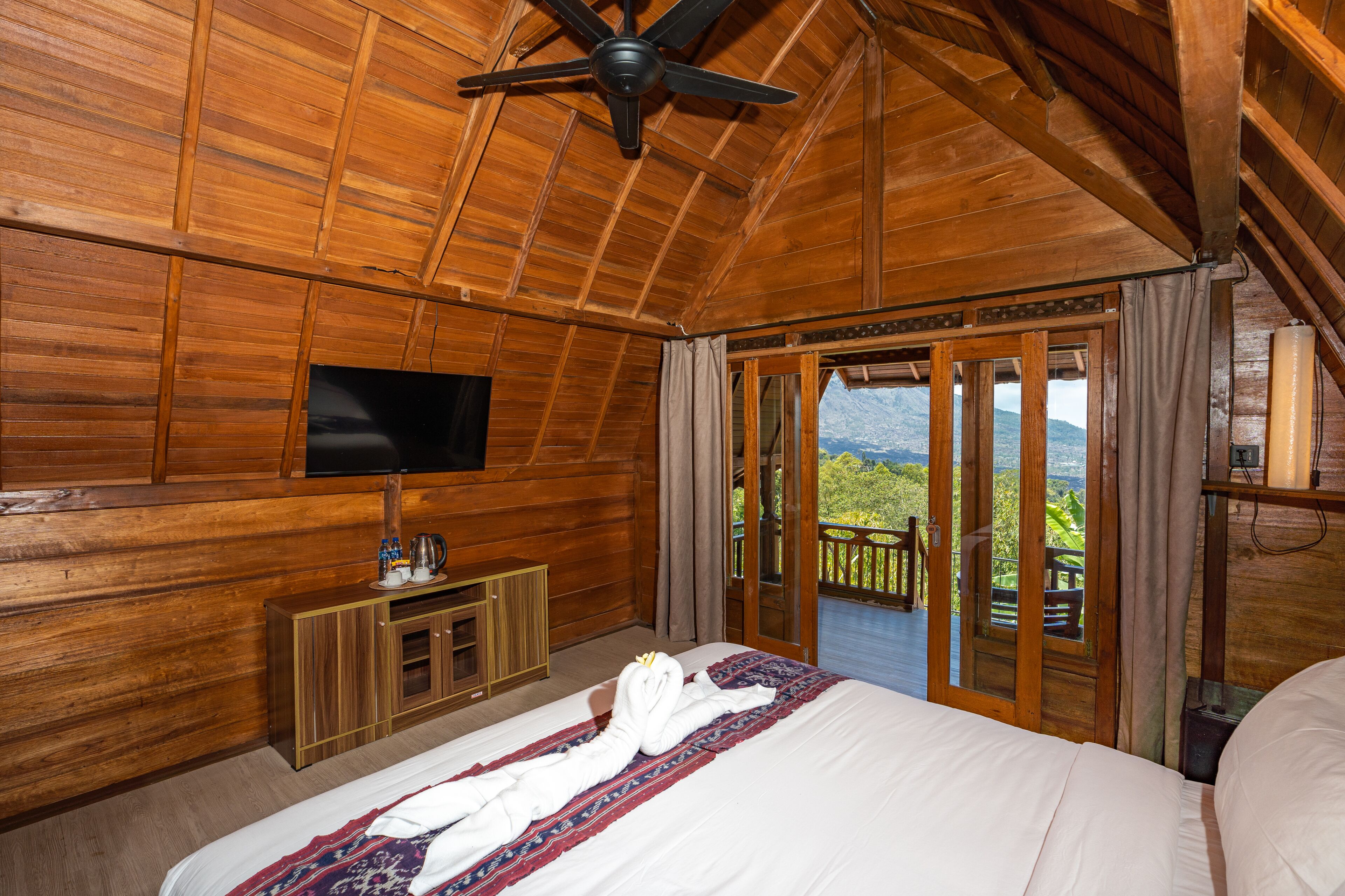 A wooden-paneled bedroom with a bed, a television, a minibar, and a balcony with a mountain view.
