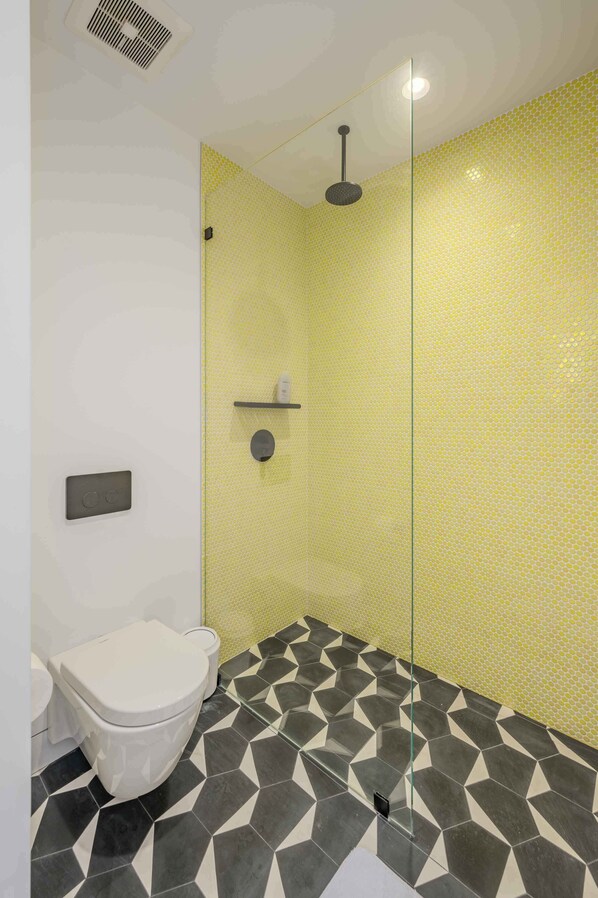 A bathroom with a white toilet, a shower with a yellow tiled wall, and a floor with black and white geometric tiles.
