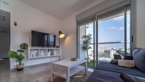 A modern living room with a large TV, a white coffee table, a dark sofa, and a view of the city through large windows. - PORT CITY HAIFA - German Colony Luxury (Haifa)