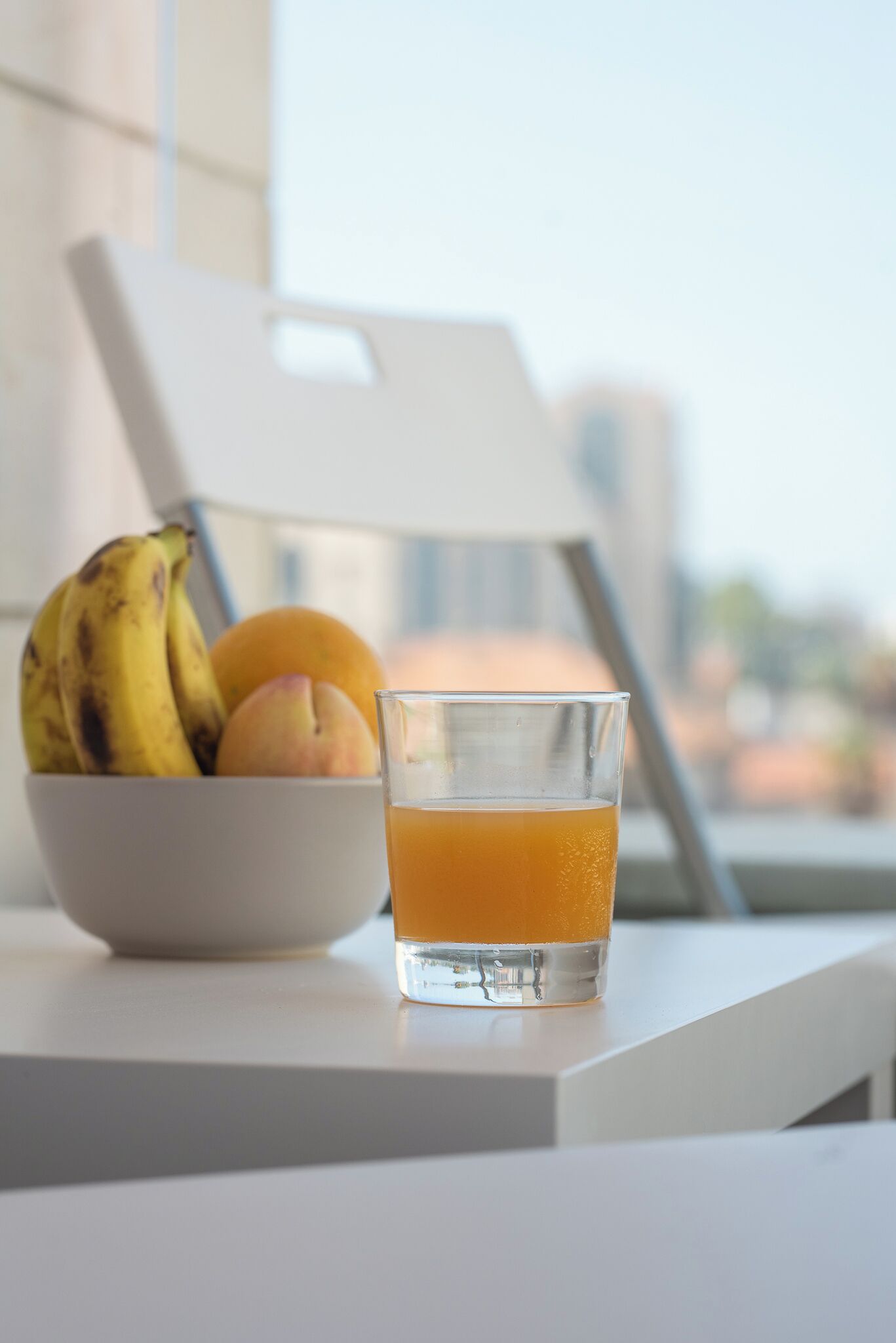 A bowl of fruit and a glass of orange juice on a table by a window.