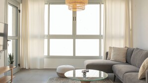 A living room with a sofa, ottoman, glass coffee table, and a hanging light fixture. - PORT CITY HAIFA - German Colony Luxury (Haifa)