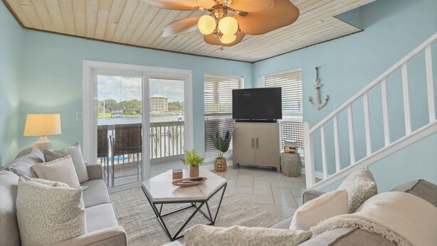 A living room with a ceiling fan, a TV on a cabinet, a sofa, and a view of a body of water through large windows. - Waterfront views! 3 BR Walk to beach, Pool, Dock (Fort Walton Beach)