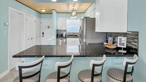 A kitchen with a black countertop, white cabinets, and a wooden ceiling. There are four chairs with cushioned seats. - Waterfront views! 3 BR Walk to beach, Pool, Dock (Fort Walton Beach)