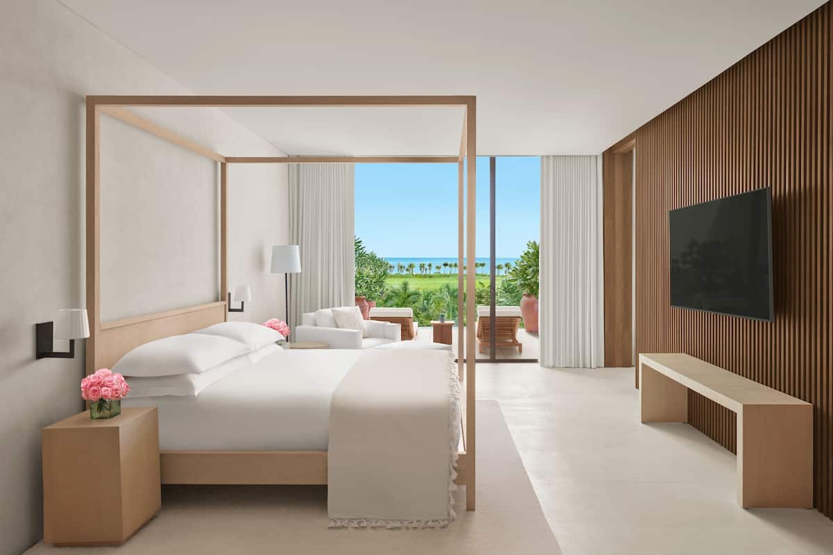 A modern hotel room with a large bed, a TV, a balcony with a view of the ocean, and a wooden wall.