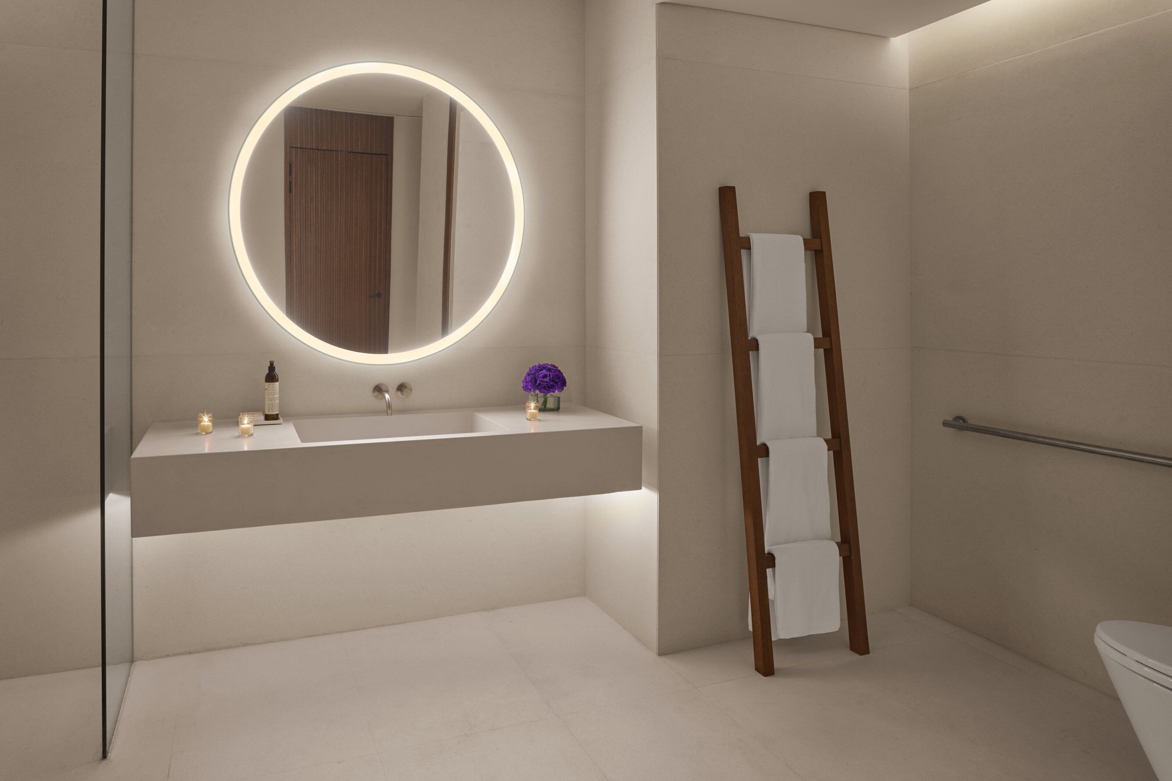 A modern bathroom with a round mirror, a wooden ladder, a towel rack, and a toilet.