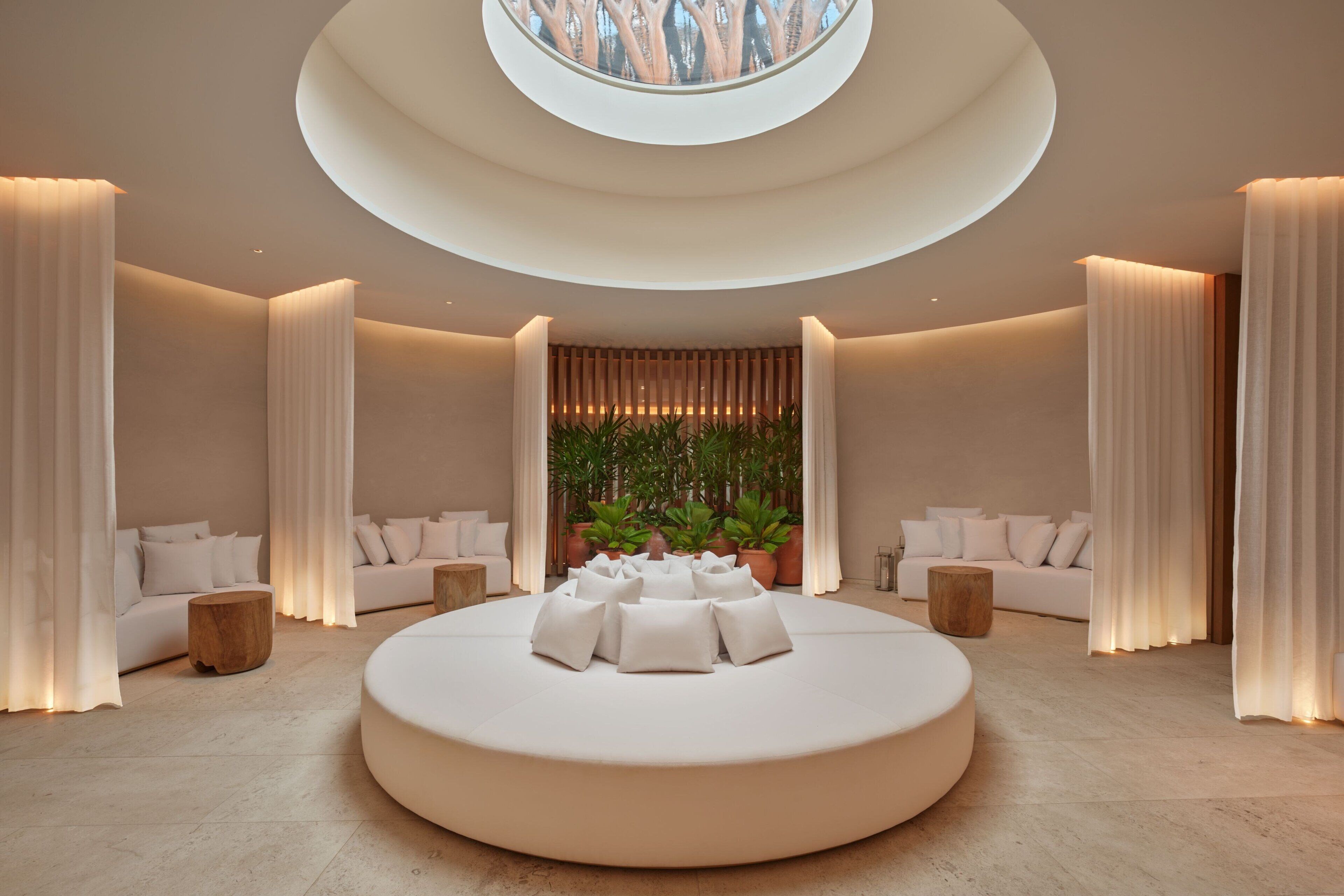 A modern, circular seating area with a central plant arrangement, surrounded by white sofas and curtains, in a well-lit room with a skylight.