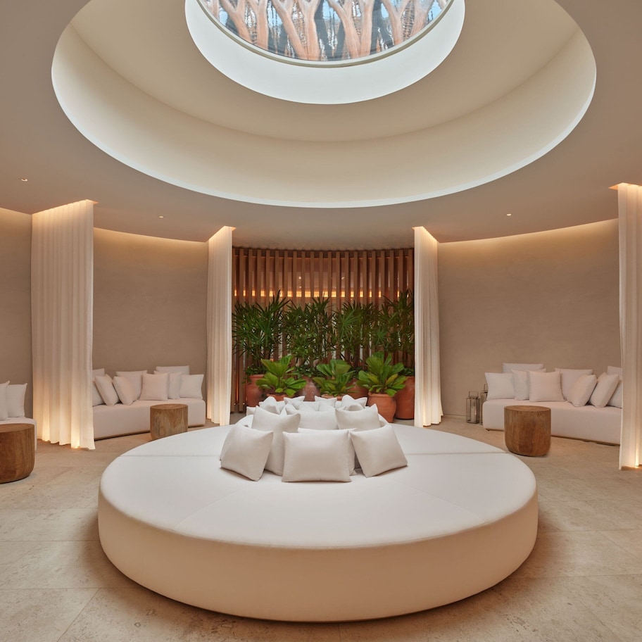 A modern, circular seating area with a central plant arrangement, surrounded by white sofas and curtains, in a well-lit room with a skylight.