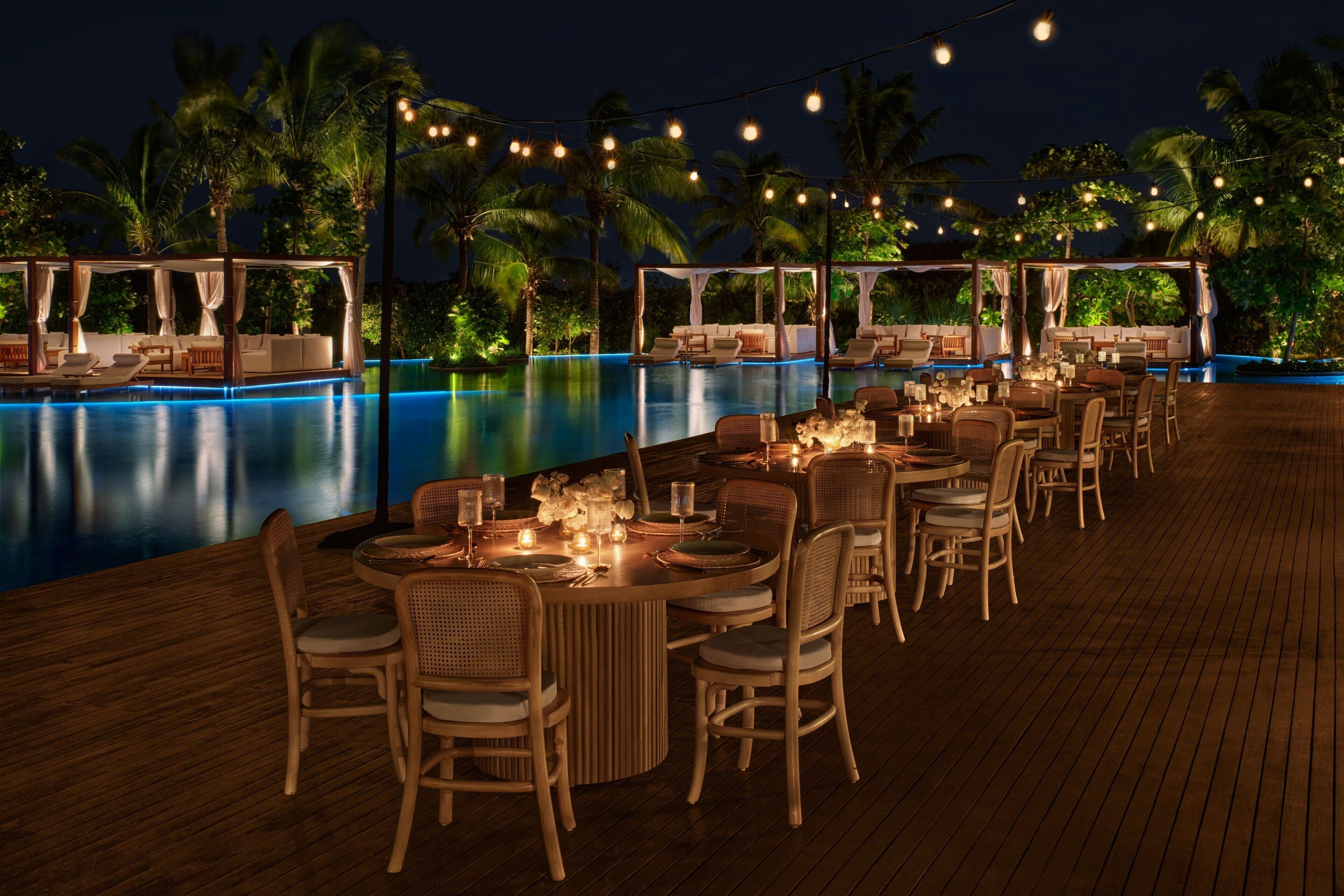 A poolside dining area with tables and chairs, string lights, and palm trees in the background.