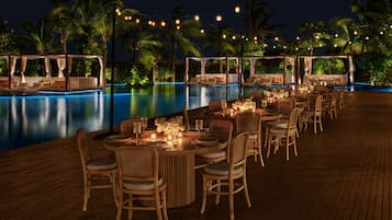 A poolside dining area with tables and chairs, string lights, and palm trees in the background.