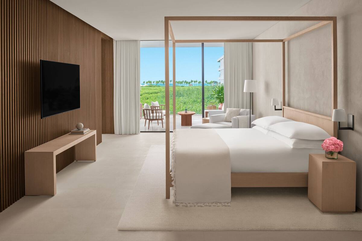 A modern hotel room with a large bed, a TV mounted on the wall, a seating area with a table and chairs, and a view of the outdoors.