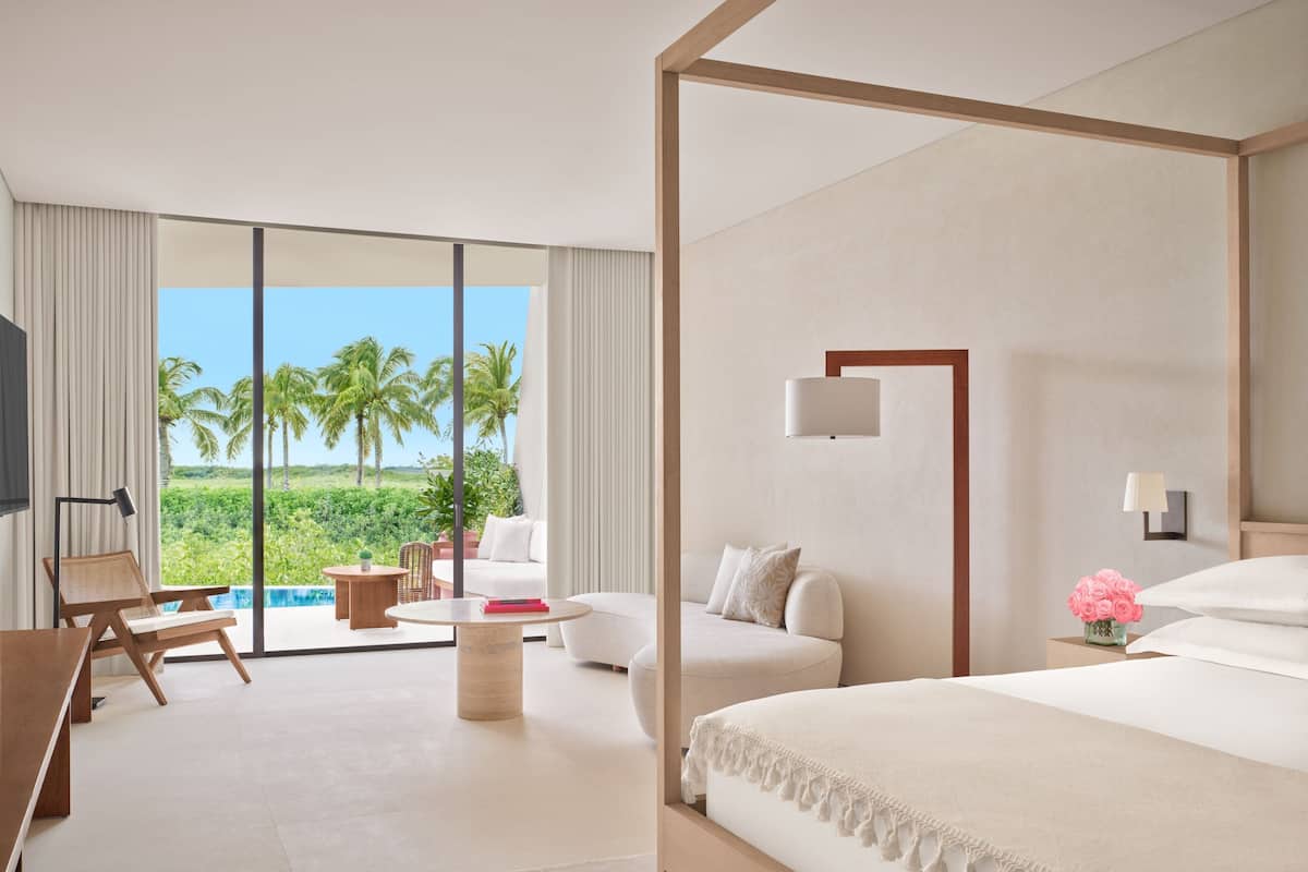 A modern hotel room with a large bed, a sofa, a desk, and a view of palm trees and a pool.