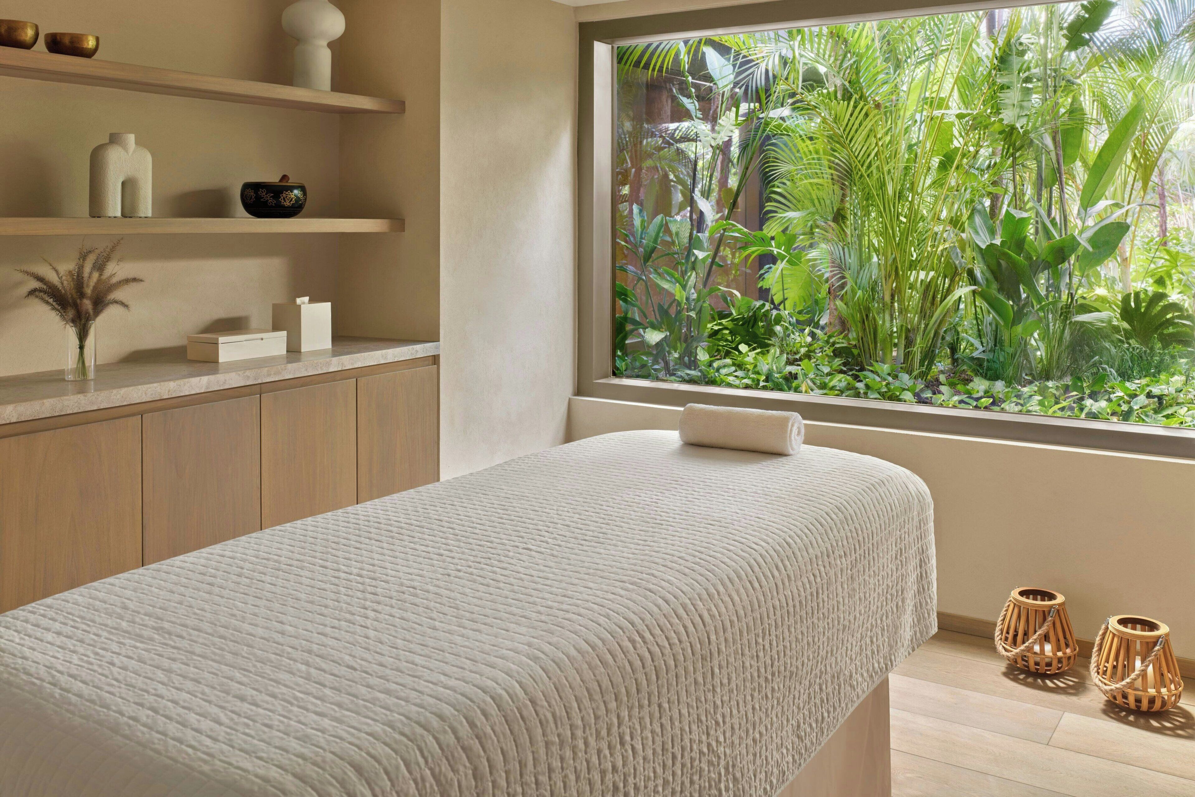A room with a bed, a window with a view of greenery, a shelf with decorative items, and a small plant on the bedside table.