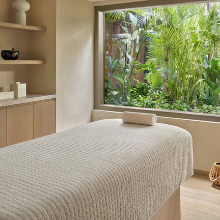 A room with a bed, a window with a view of greenery, a shelf with decorative items, and a small plant on the bedside table.