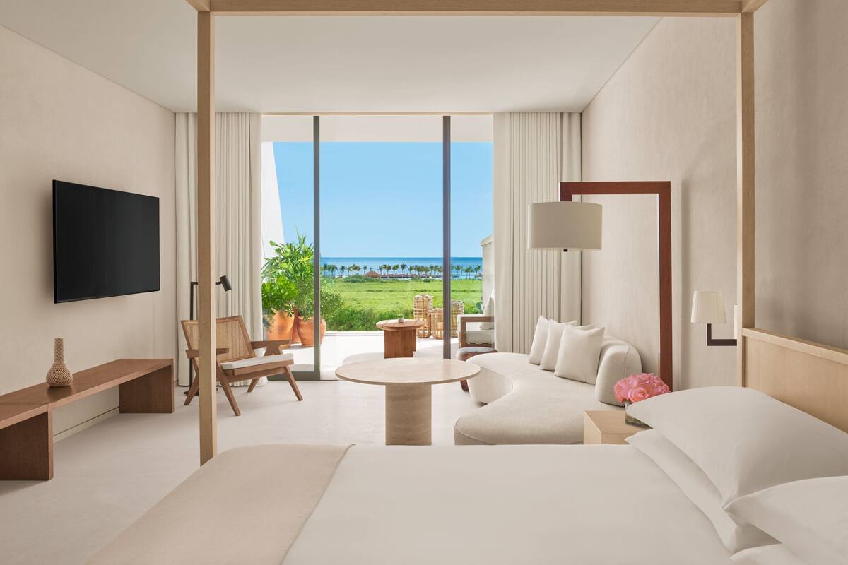 A modern hotel room with a large bed, a sofa, a TV, and a balcony with a view of the ocean.