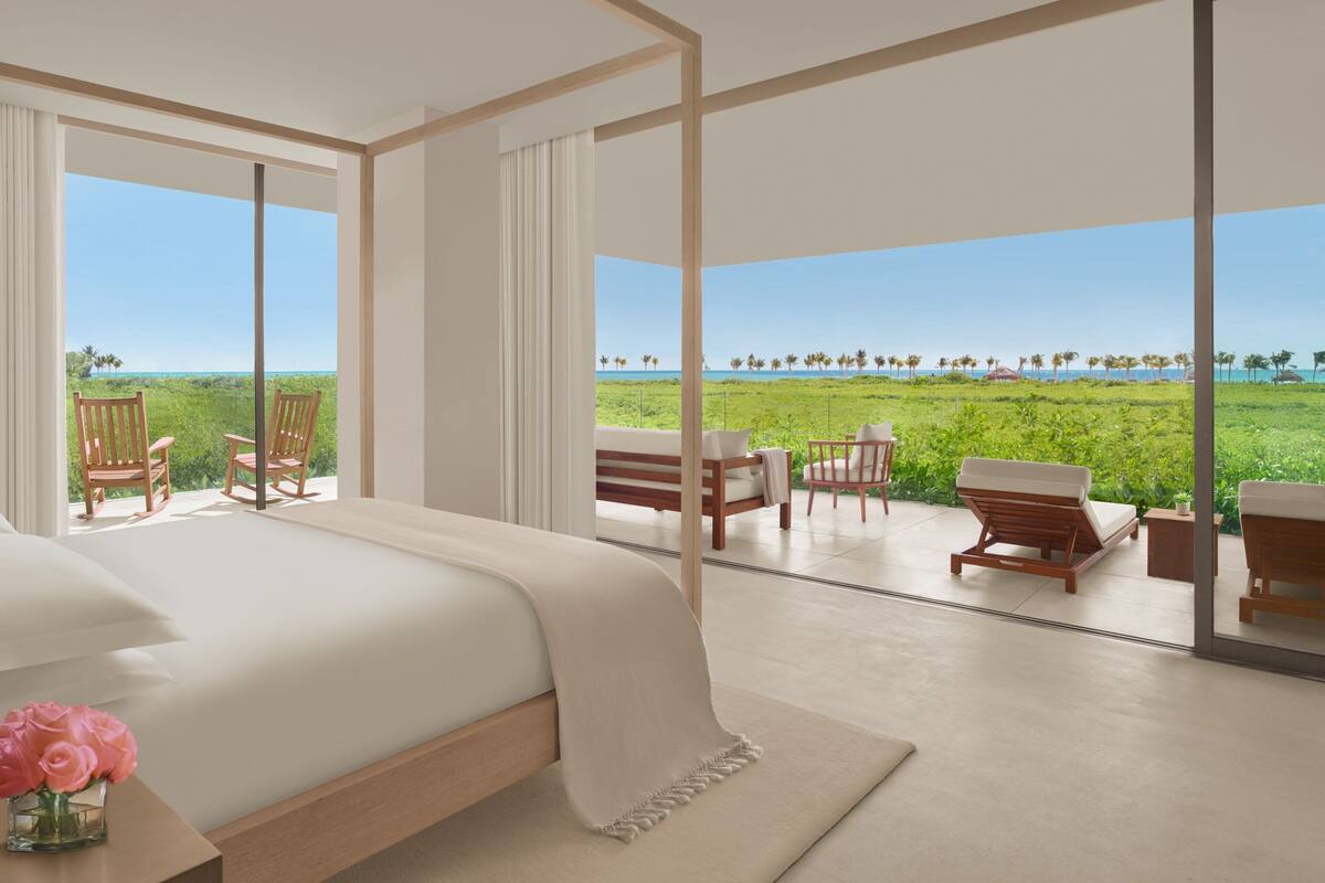 A bedroom with a large bed, a nightstand with a vase of flowers, and a view of a green landscape through large windows.