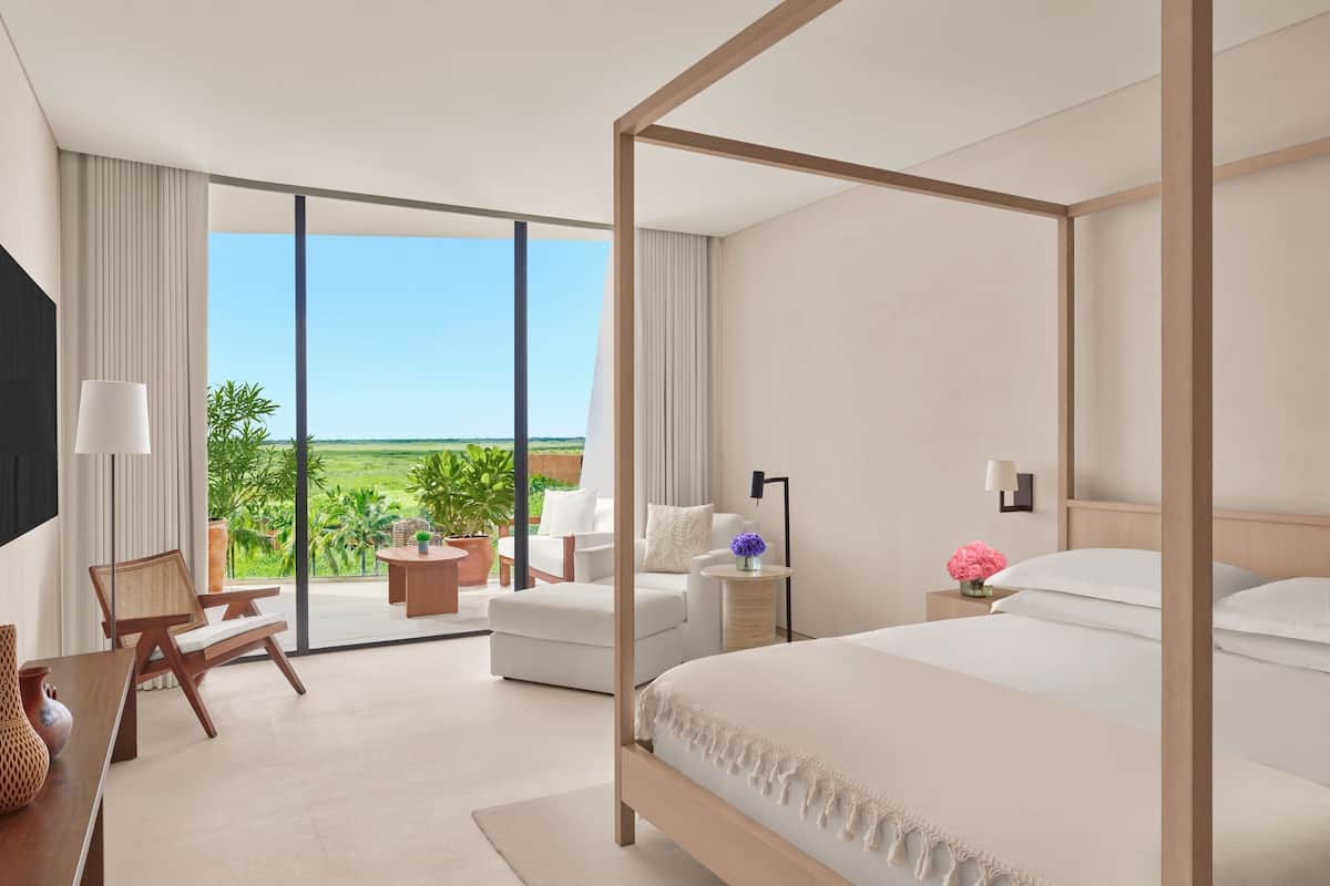 A modern hotel room with a large bed, a balcony with seating, and a view of the outdoors.