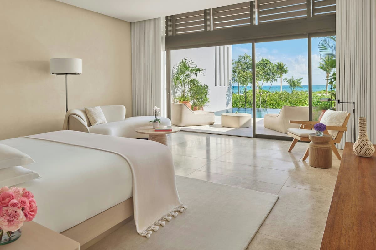 A modern hotel room with a large bed, two armchairs, a small table, and a view of the ocean.