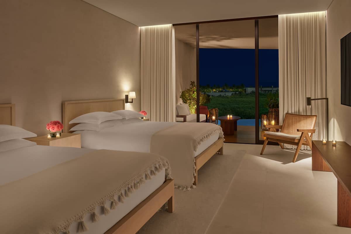 A hotel room with two beds, a seating area with a chair and table, and a view of the outdoors.