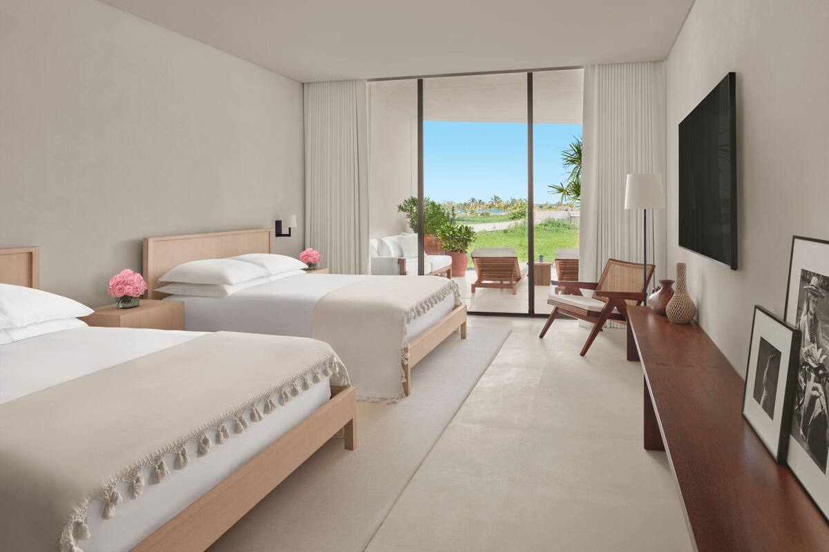 A hotel room with two beds, a desk, a chair, a TV, and a balcony with a view of the beach.
