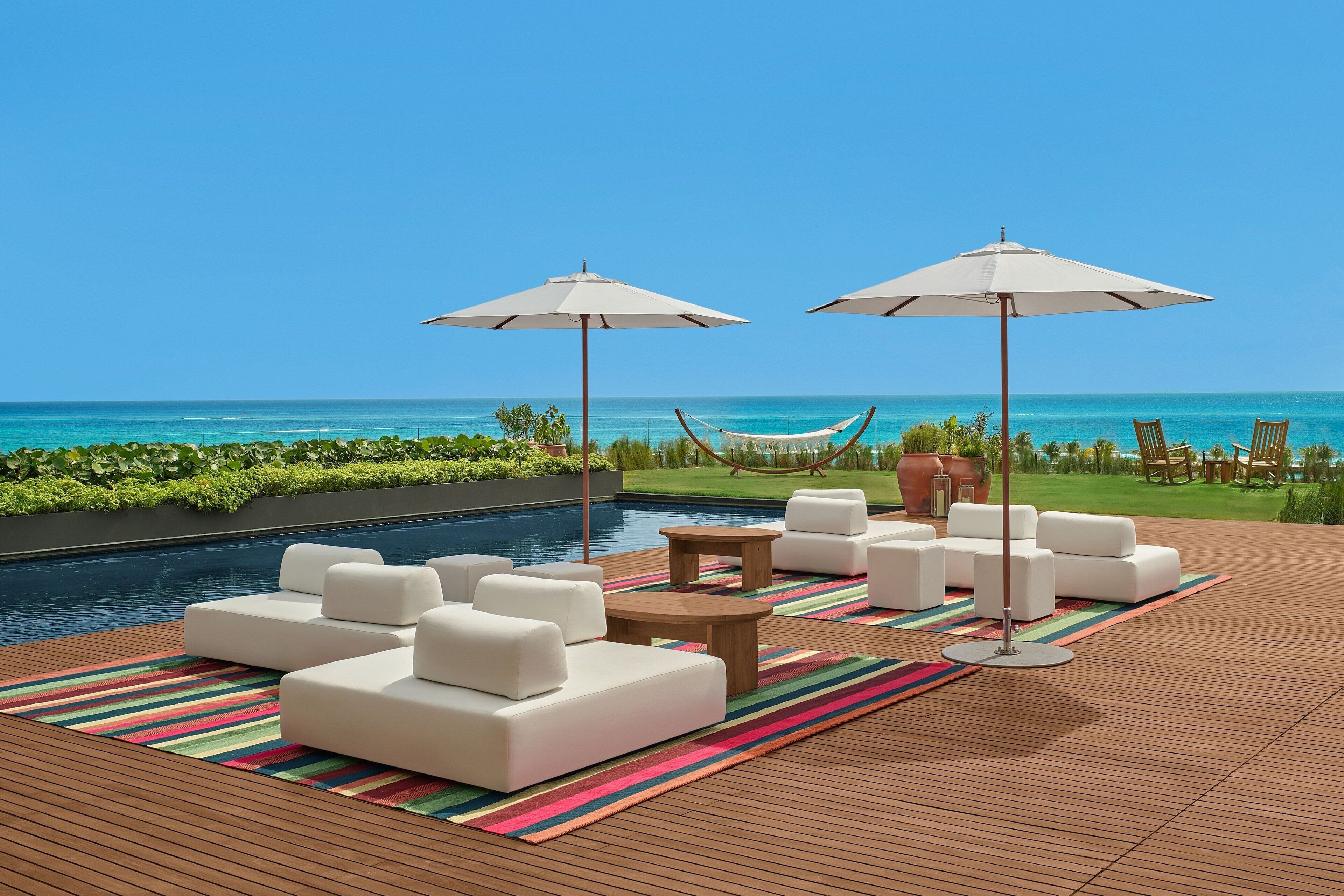 A poolside area with white sofas, two umbrellas, a hammock, and a view of the ocean.