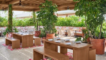 Een buitenruimte met eettafels en banken van hout, omringd door potplanten en een betegelde vloer.