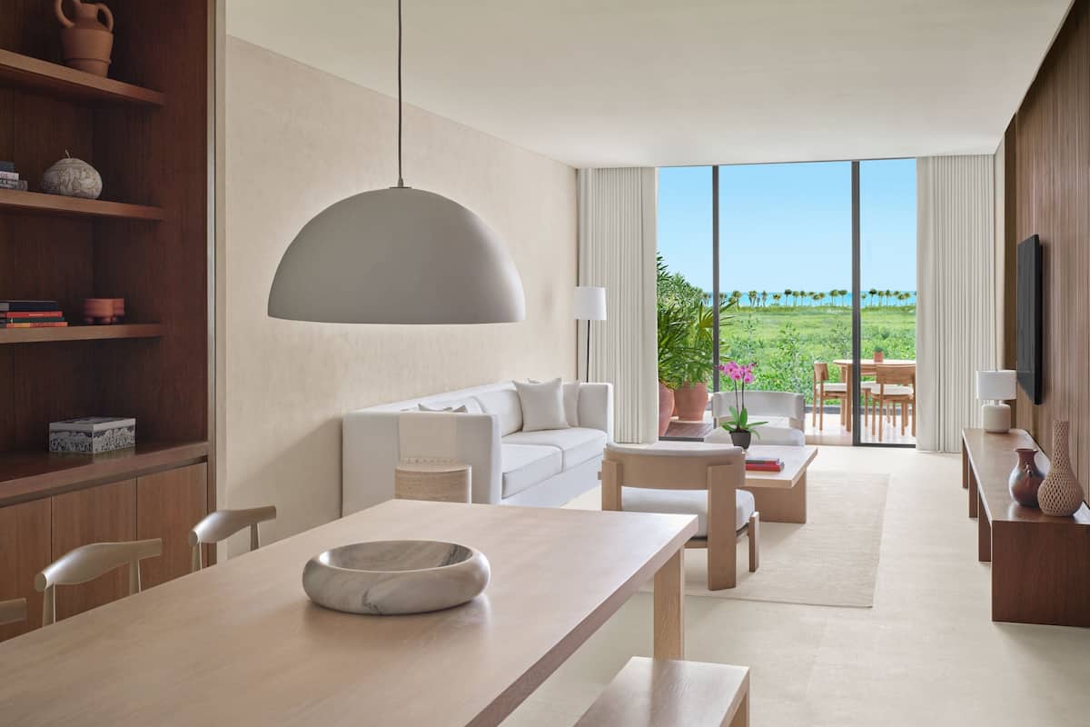 A modern living room with a sofa, dining table, and a large window offering a view of greenery and a clear sky.