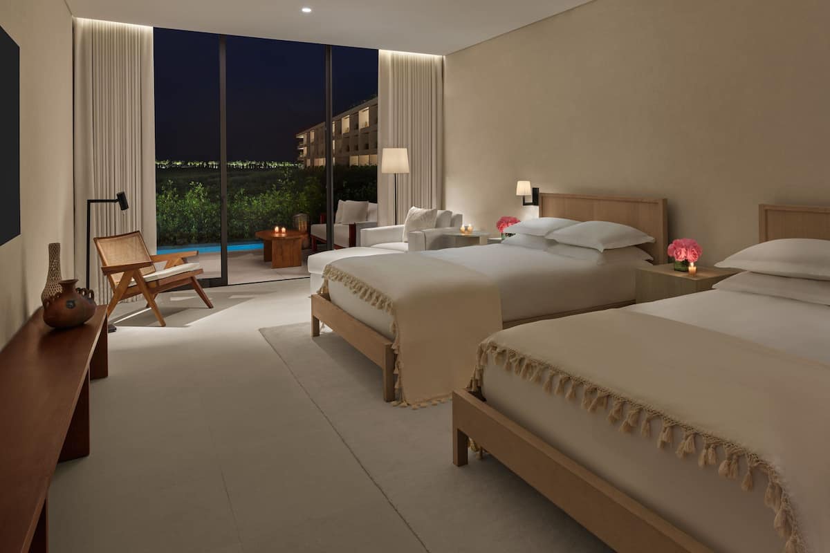 A hotel room with two beds, a seating area with a chair and a table, and a view of the outdoors.