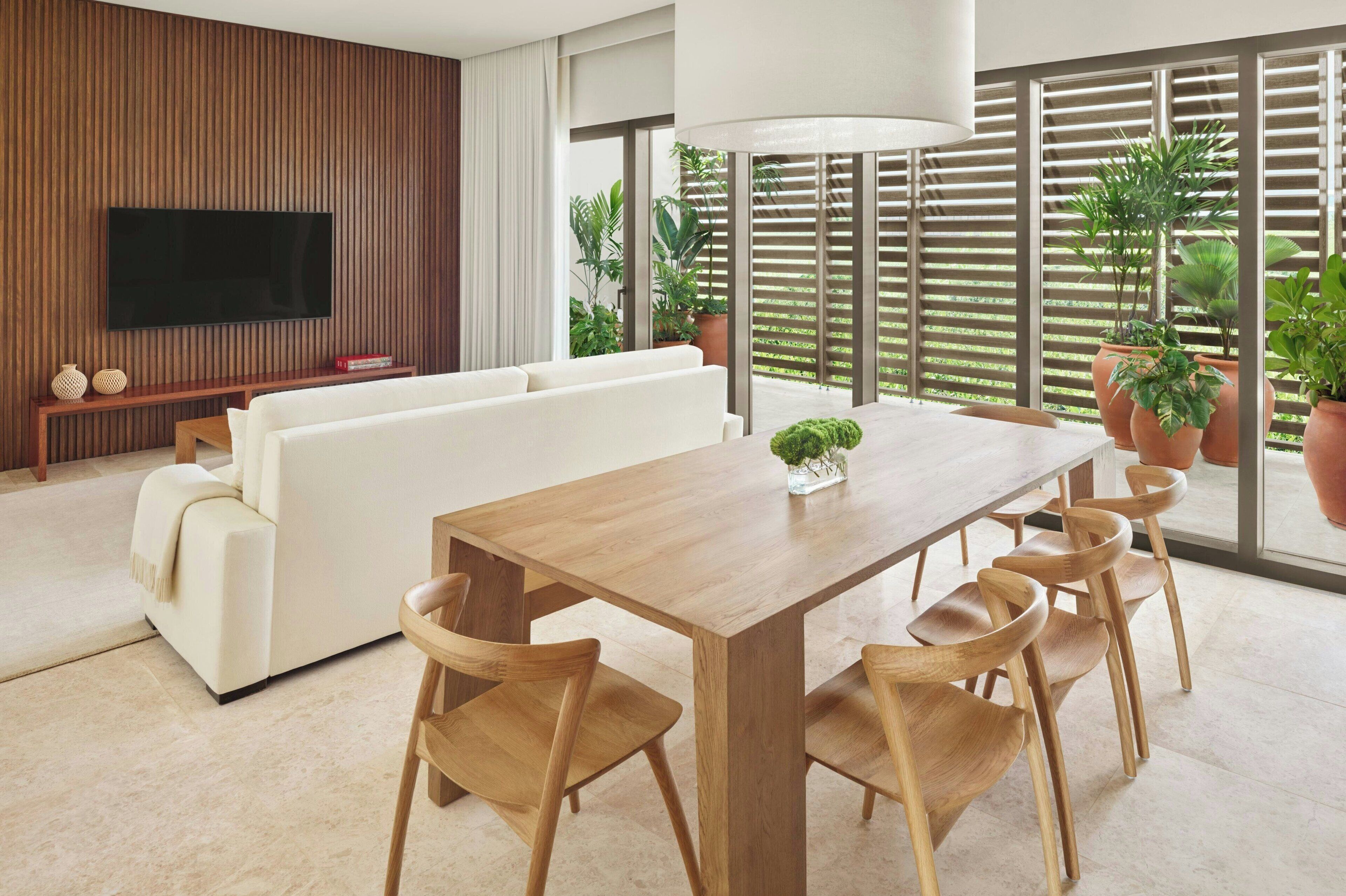 Un salon moderne avec un lambris en bois, un téléviseur à écran plat, un canapé blanc, une table à manger en bois avec des chaises, et de grandes fenêtres ornées de plantes en pot.